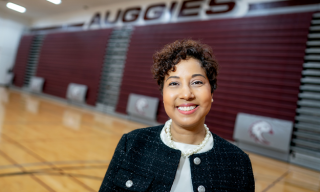 A person smiling in a gymnasium with maroon bleachers and "AUGGIES" sign in the background.