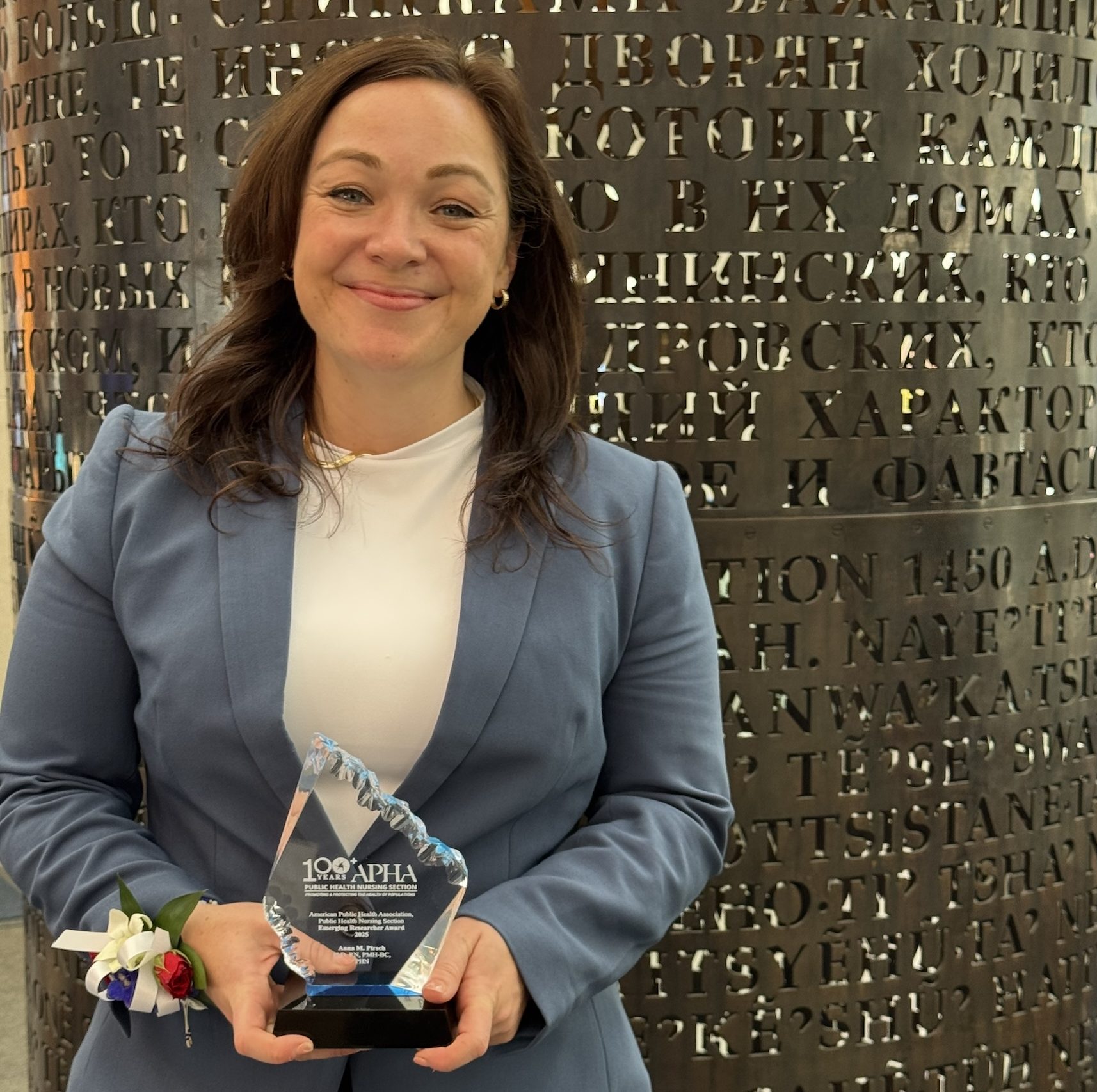 Anna Pirsch stands in front of a bronze column holding a clear award plaque. She is wearing a blue blazer, a white blouse, and flowers on her wrist.