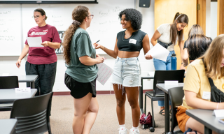People interacting in a classroom setting, holding papers and conversing.