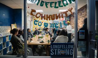 A community meeting visible through a window with "LONGFELLOW COMMUNITY COUNCIL" written on it, featuring people sitting around a table.