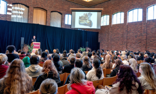 A lecture in a large brick-walled auditorium with a speaker at a podium and attendees seated.