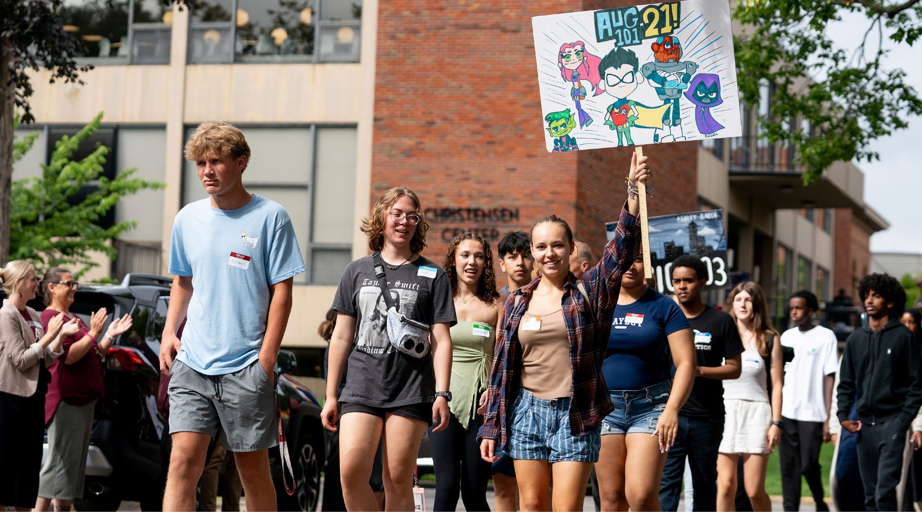 A group of young people walking in front of a building, one holding a sign with cartoon superheroes and the date "AUG 10! 21!"