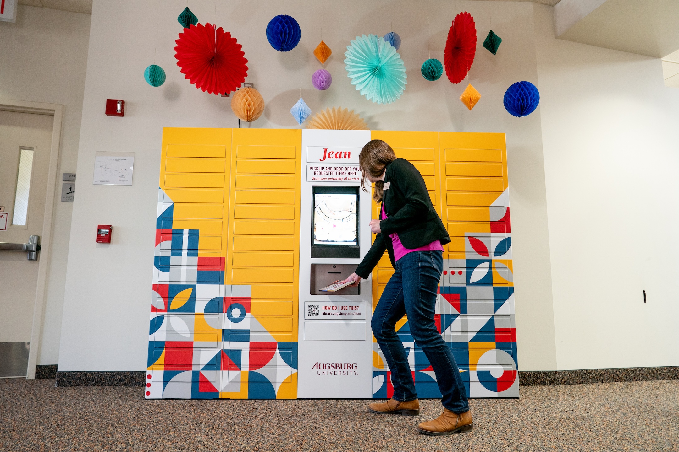A librarian retrieves a book from "Jean," a brilliant yellow smart locker system with multicolored designs and paper decorations hanging above it.