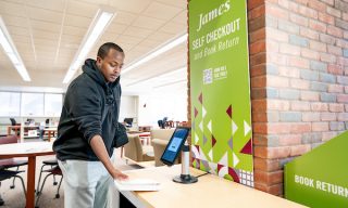 A student checks out a book at Lindell Library below a green sign that reads "James, Self Checkout"