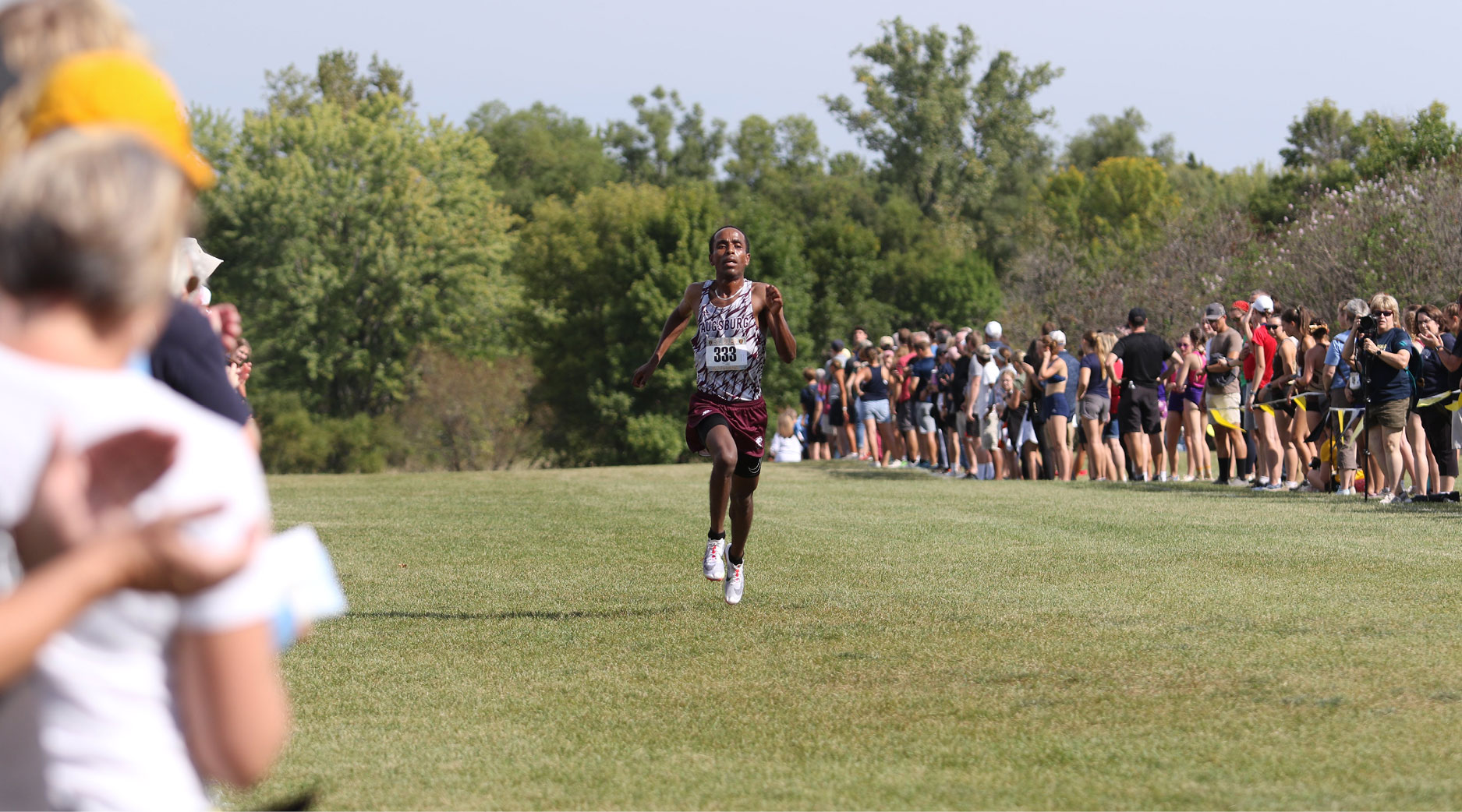 A male cross country runner wearing a maroon and white patterned singlet with the number 330 sprints across a grass field toward the finish line, while spectators line the course on the right.