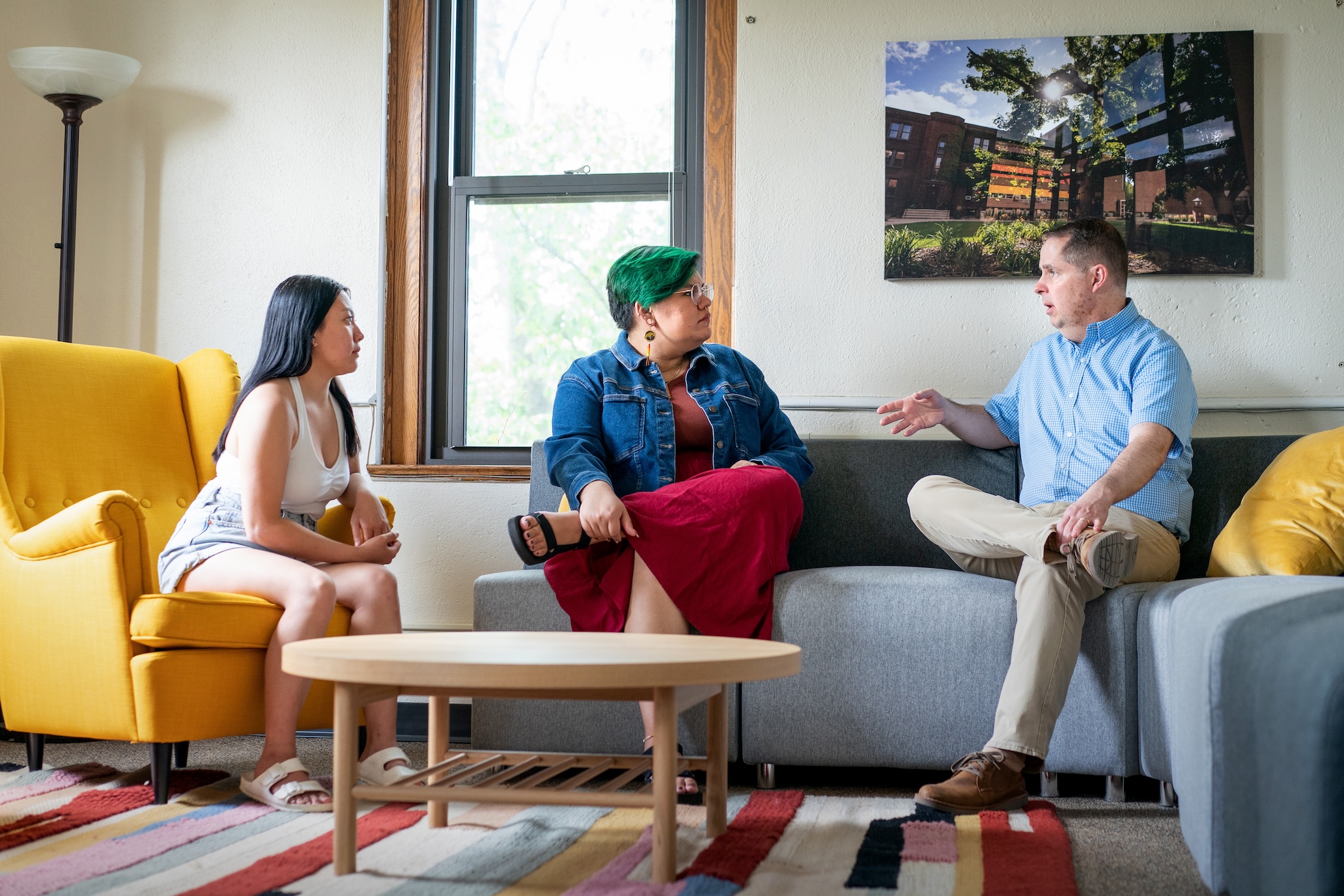 Professor Tim Pippert and two Augsburg Family Scholars sit on gray and yellow furniture having a conversation in the AFS lounge