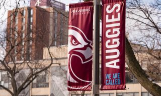 Maroon flagpole banners with the Auggie Eagle and text reading We Are Called Auggies against a backdrop of campus buildings
