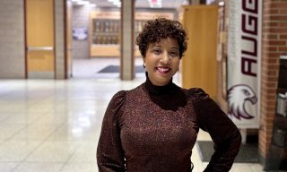 Woman in a sparkly brown turtleneck and dark pants posing in a brightly lit hallway with a banner that reads "AUGGIES."
