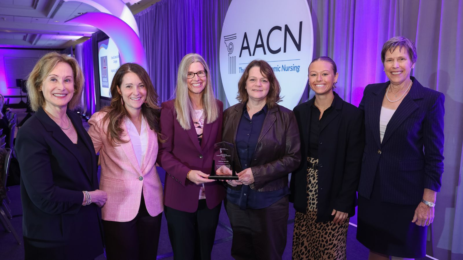 Six individuals wearing blazers and dress clothes stand in a row and smile. The person in the middle holds a clear award. The AACN is printed on a screen behind them.