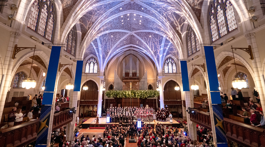A wide-angle interior view of a large, high-vaulted Gothic-style church during a performance, likely Christmas or Advent. A choir and orchestra are on a raised platform near the front, beneath a large pipe organ and festive green wreaths. The church is filled with an audience, and blue banners hang from the balconies on either side.