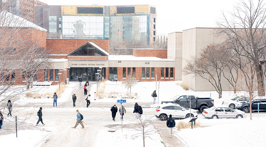 A wide shot of the Foss Center on a college campus, covered in snow. Several people are walking on the snowy paths and sidewalk in front of the building, and parked cars are visible on the right.