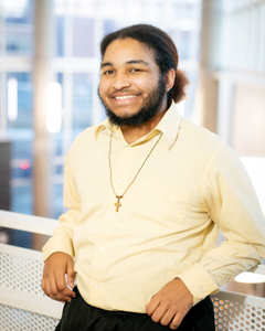 A headshot of a smiling young man in a light yellow button-down shirt leaning against a white railing indoors.