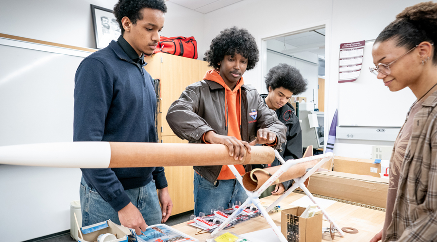 Four students working together in a physics lab to assemble a large model rocket on a white PVC pipe stand.