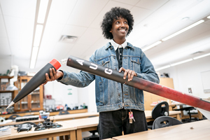 A smiling student in a denim jacket holding a black and red model rocket, detaching the nose cone in a science lab setting.