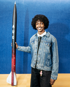 A portrait of a smiling student in a denim jacket standing next to a tall, black and red model rocket labeled "AUG."