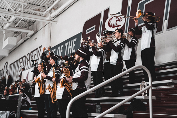 A college pep band in maroon and white uniforms plays brass and saxophone instruments from the bleachers during a hockey game.