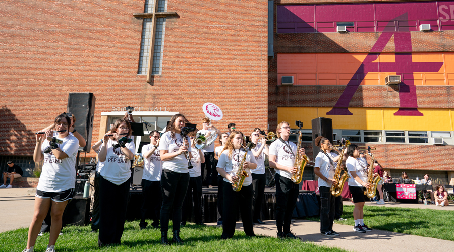 A pep band in white t-shirts performs outdoors on a lawn in front of "Science Hall" during a Homecoming event.