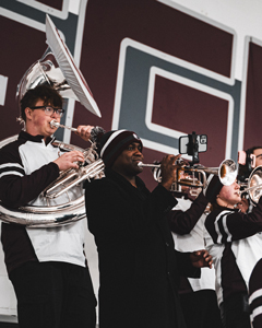 A close-up of three pep band members playing a sousaphone and trumpets against a wall with large maroon graphic lettering.
