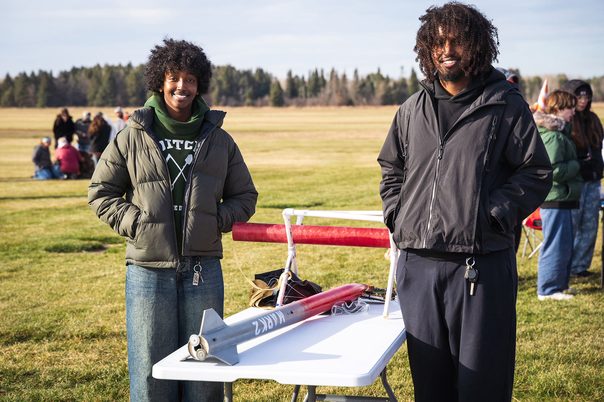 SoNSRockets-104 Two smiling young men stand behind a white folding table in a field, showcasing their "MARK 2" model rocket.