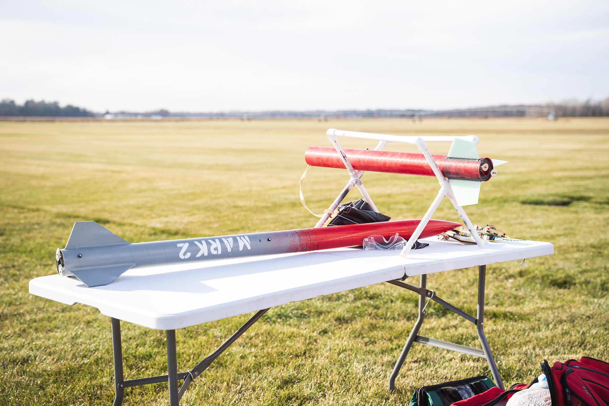 SoNSRockets-108 A "MARK 2" model rocket and another red rocket body rest on a white folding table in an open, grassy field.