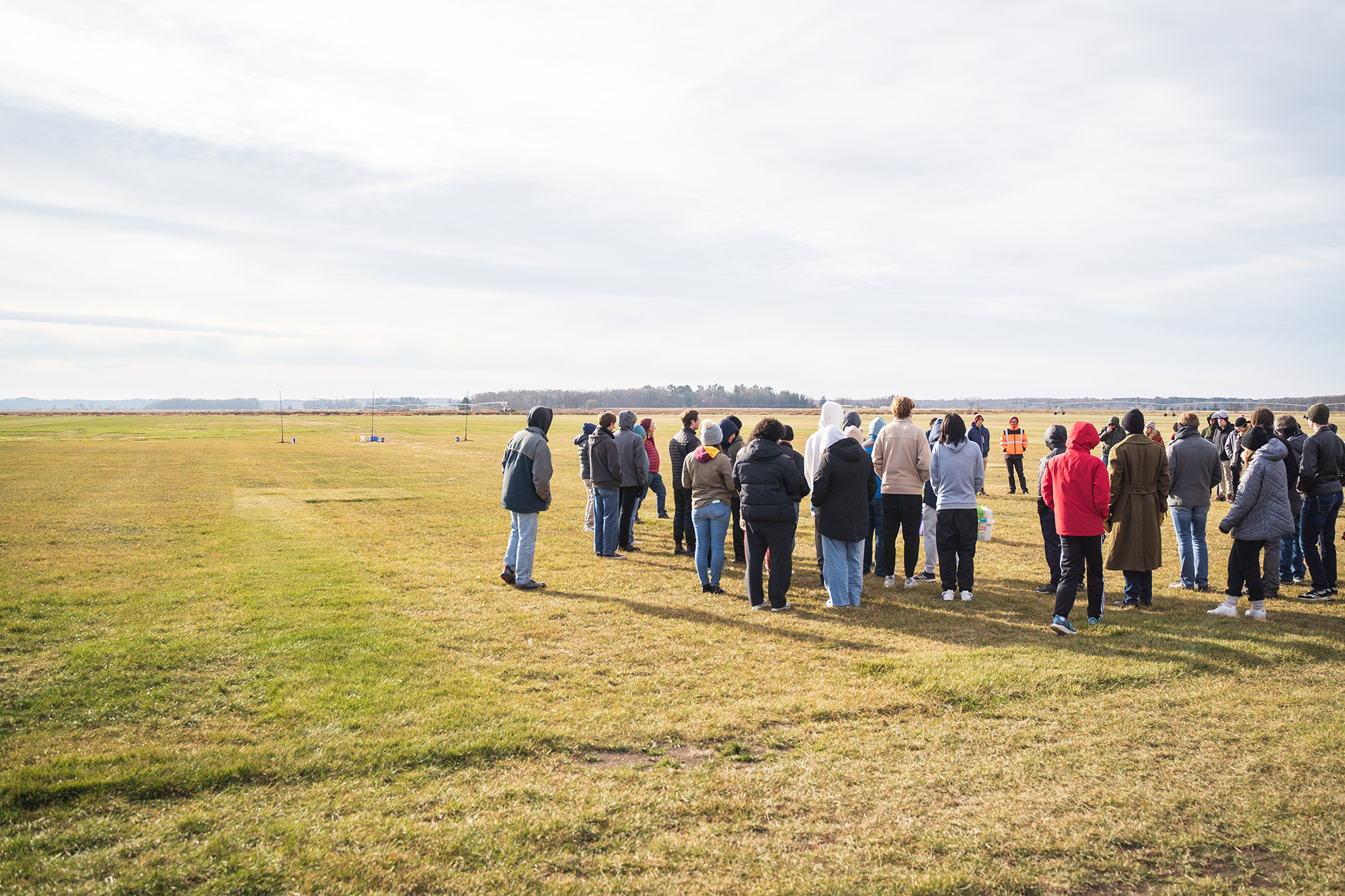 SoNSRockets-110 A large group of people gather in a wide, grassy field for a briefing or meeting under a bright, overcast sky.