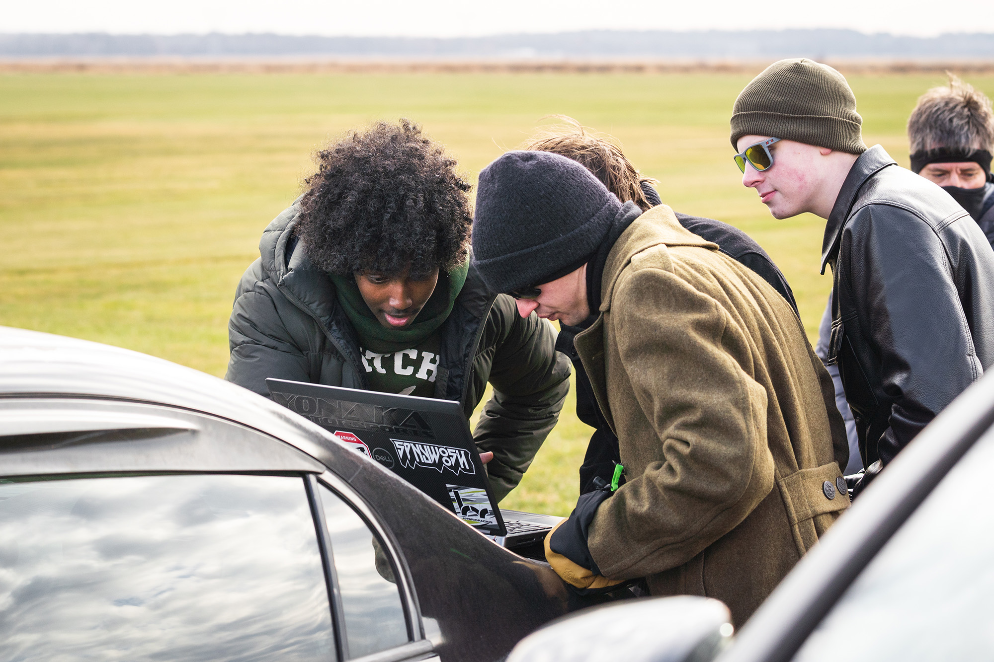 SoNSRockets-111 A group of people huddle together outdoors to look at a laptop screen resting on the trunk of a car.