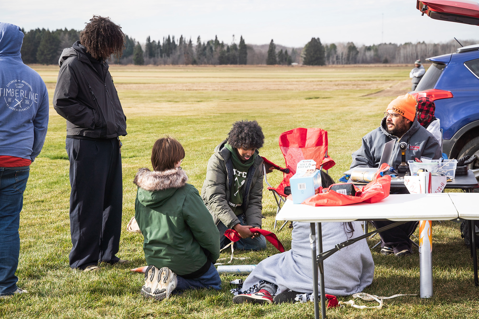SoNSRockets-116 A group of people sit and stand around a field workspace, assembling parts and preparing equipment for a rocket launch.