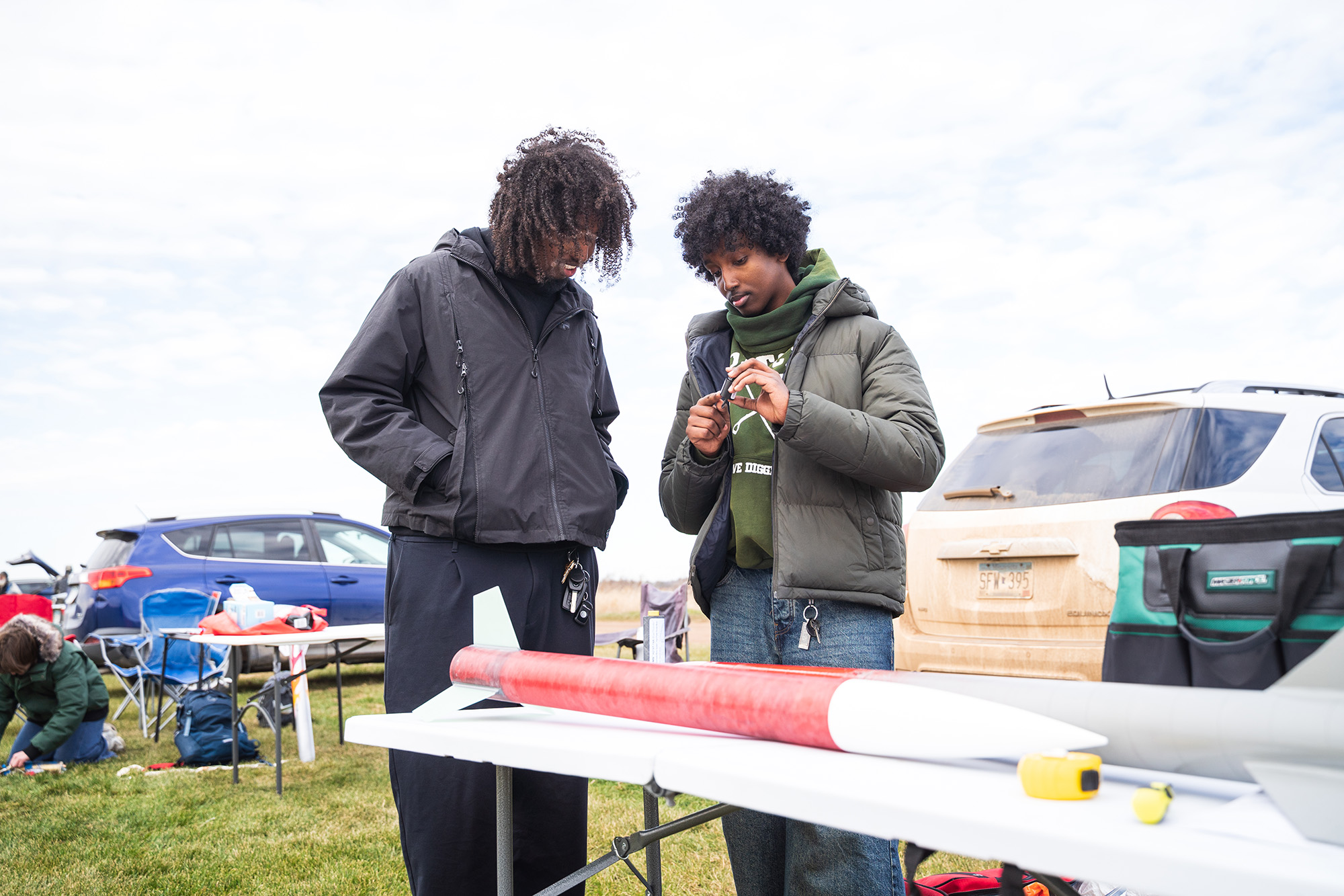 SoNSRockets-122 Two people stand behind a table in a field, closely examining a small rocket component held in their hands.