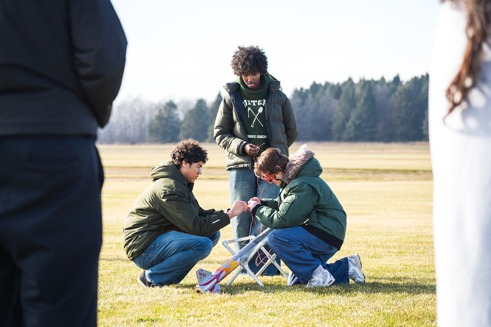 SoNSRockets-135 Three people kneel in a field to carefully connect wiring to a silver model rocket decorated with flame decals.