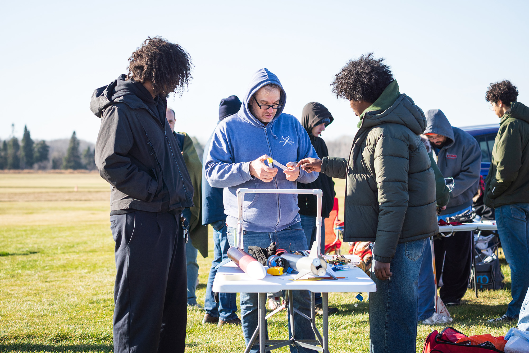 SoNSRockets-138 A group of people gather around a table in a field to inspect components for model rockets.