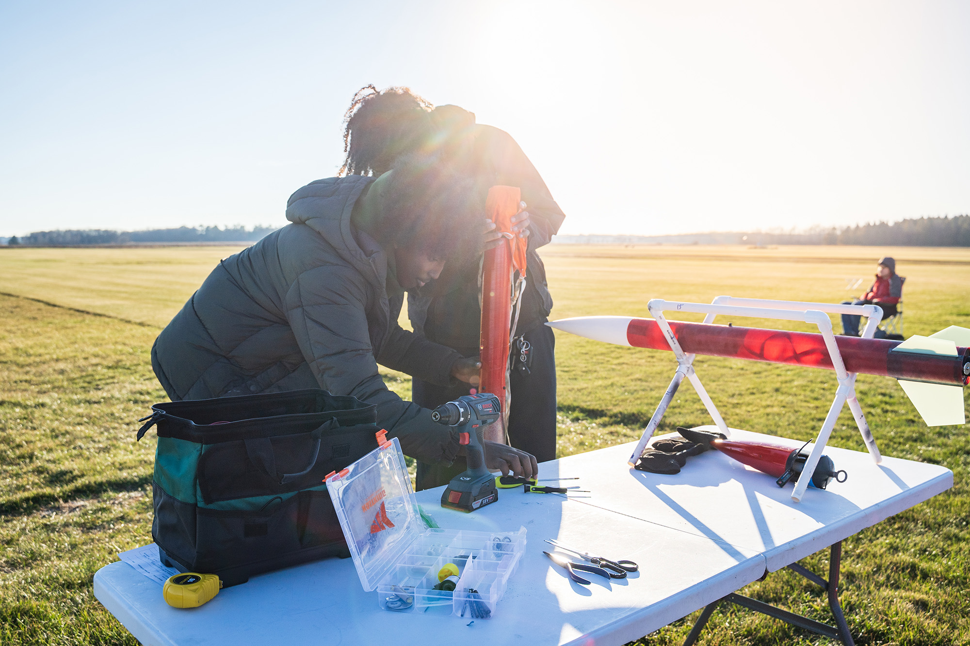 SoNSRockets-156 Two people work on a large red model rocket at a table filled with tools, including a drill and a hardware organizer, in a sunlit field.