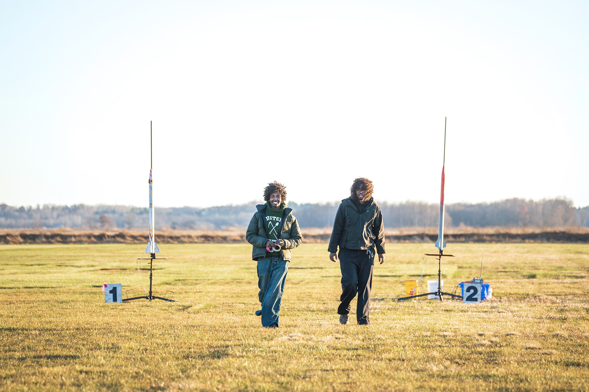 SoNSRockets-176 Two young men walk across an open field toward the camera, flanked by two large model rockets mounted on launch stands.