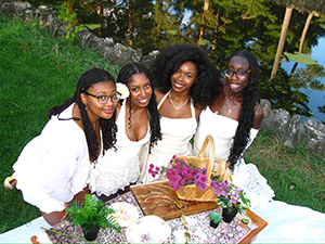 Four Black women wearing white dresses pose for a photo at an outdoor picnic near a body of water.
