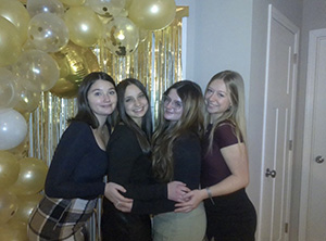 Gemini said Four young women posing and smiling together in front of a celebratory backdrop of gold and white balloons and a gold tinsel curtain.