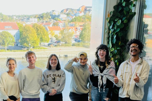 A group of six diverse students smiling and posing together indoors in front of a large window overlooking a coastal town.