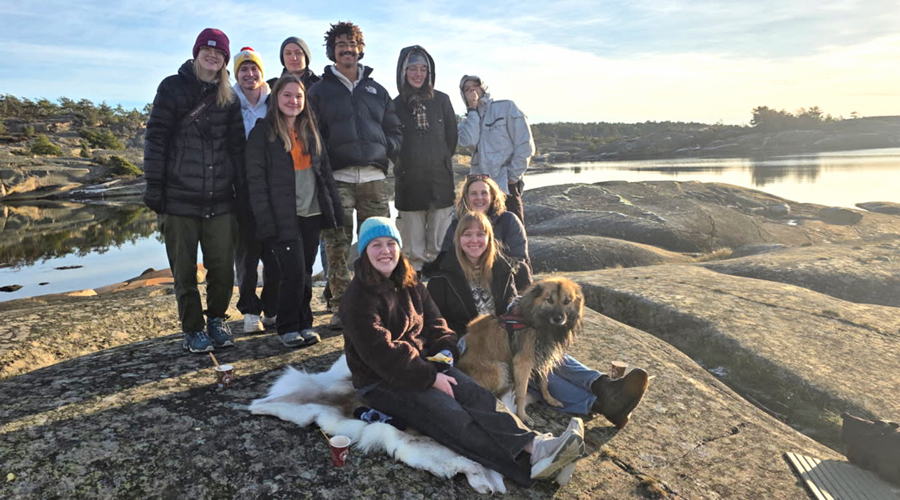 A group of students in winter gear posing on a rocky coastline next to a calm body of water at sunset.