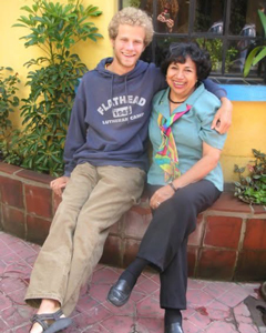 A young man in a blue hoodie and an older woman in a teal blouse sitting together and smiling on a brick ledge.