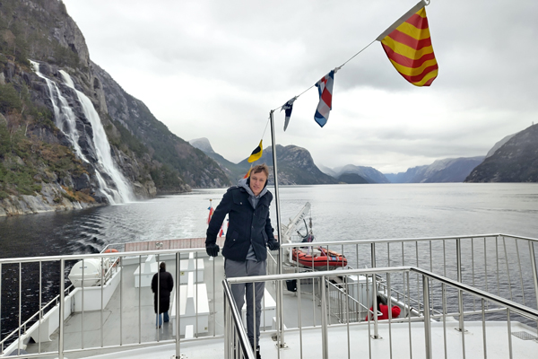 A man standing on the deck of a boat in a Norwegian fjord, with a large waterfall and misty mountains in the background.