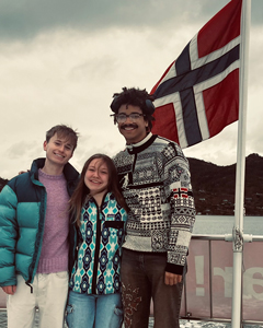 Three students smiling on a boat deck with a Norwegian flag waving in the background.
