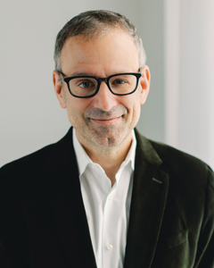 A professional headshot of a man with glasses and a slight smile, wearing a dark velvet blazer over a white shirt.
