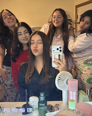 Five young women pose for a mirror selfie in a bedroom; the woman in front makes a kissing face while the others smile and lean in.