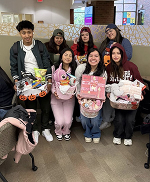 A group of seven students posing together indoors, with four of them holding large gift baskets filled with snacks and stuffed animals.