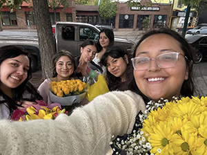 A group of six young women taking a selfie outdoors on a city sidewalk while holding several bouquets of bright yellow flowers.