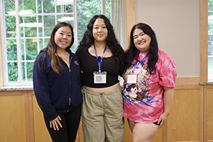 Three young Hmong women posing and smiling together indoors, wearing event lanyards.
