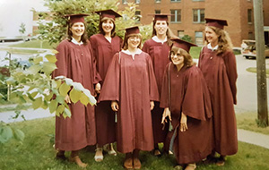 Six women in maroon graduation caps and gowns posing together on a grassy campus lawn.