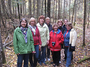 A group of eight women standing together on a wooded trail during autumn.