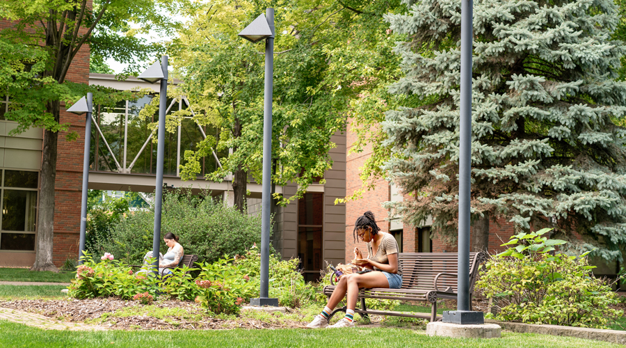Two students sit on separate park benches in a lush, green campus courtyard. Modern black lamp posts and a brick building with a glass walkway are visible behind them under the shade of mature trees.