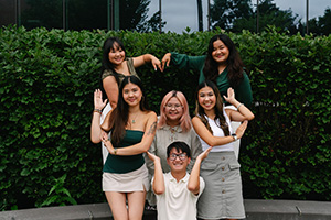 Six Asian American students posing playfully in front of a green hedge, with two making a heart shape with their hands.