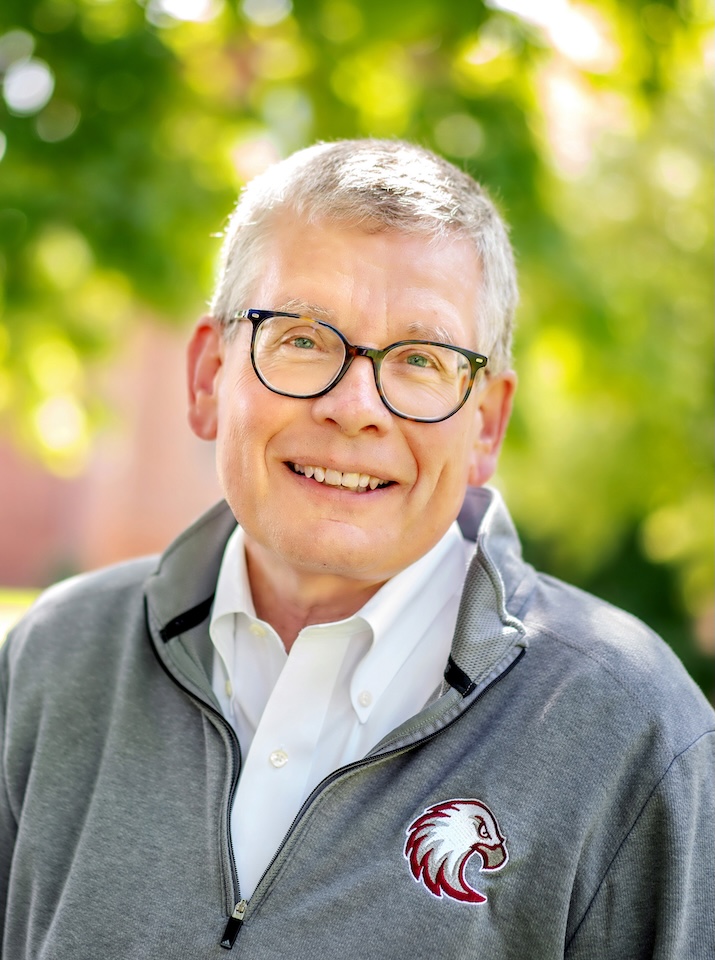 Paul Pribbenow smiles at the camera against a background of foliage. He is wearing a white collared shirt and a gray Augsburg fleece.