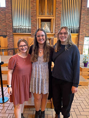 Three young women smiling together in front of a large pipe organ inside a church or chapel.